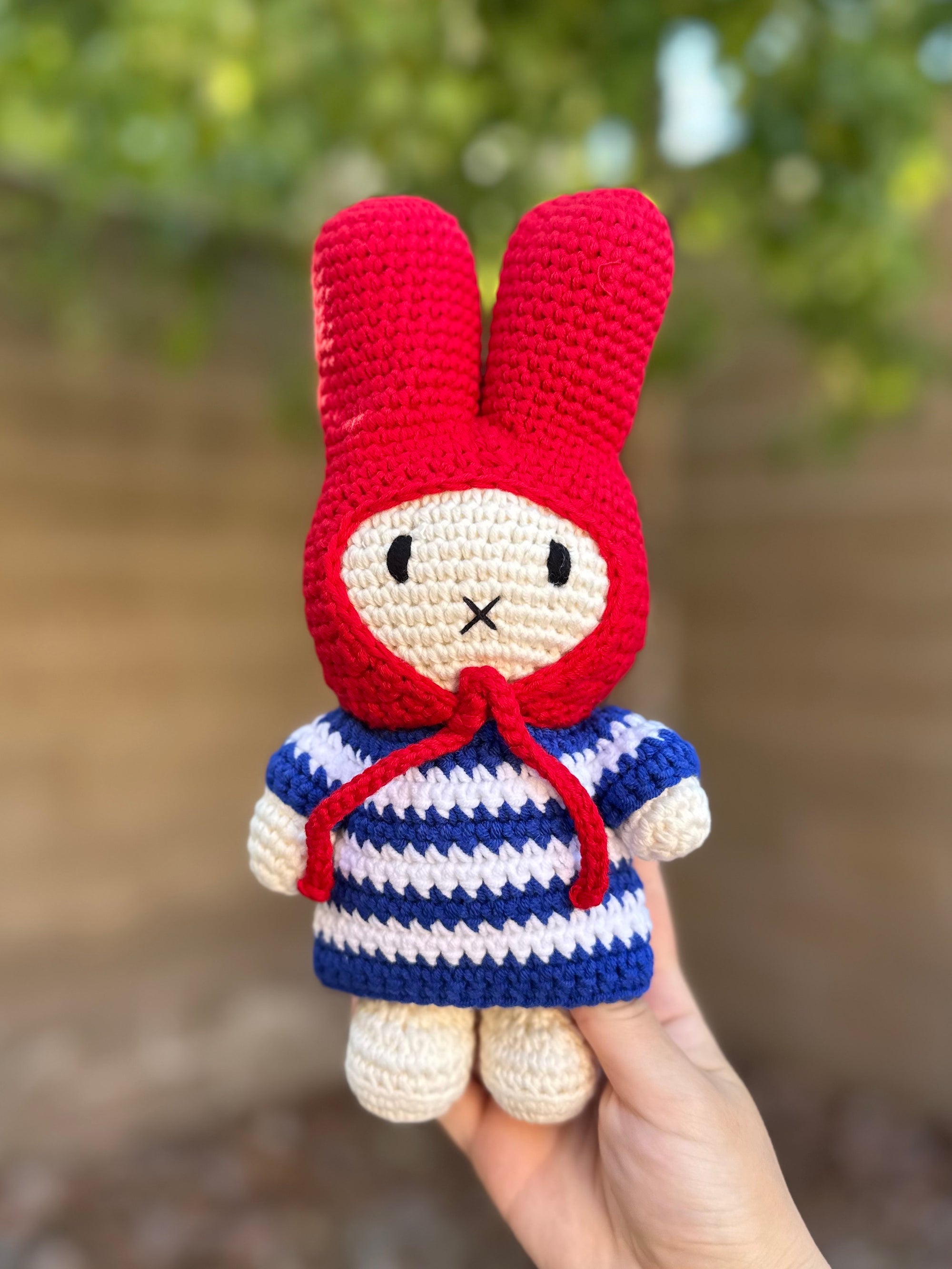 Handmade Miffy crochet plush in a blue-and-white striped dress with a bright red bonnet, held outdoors against a blurred background.
