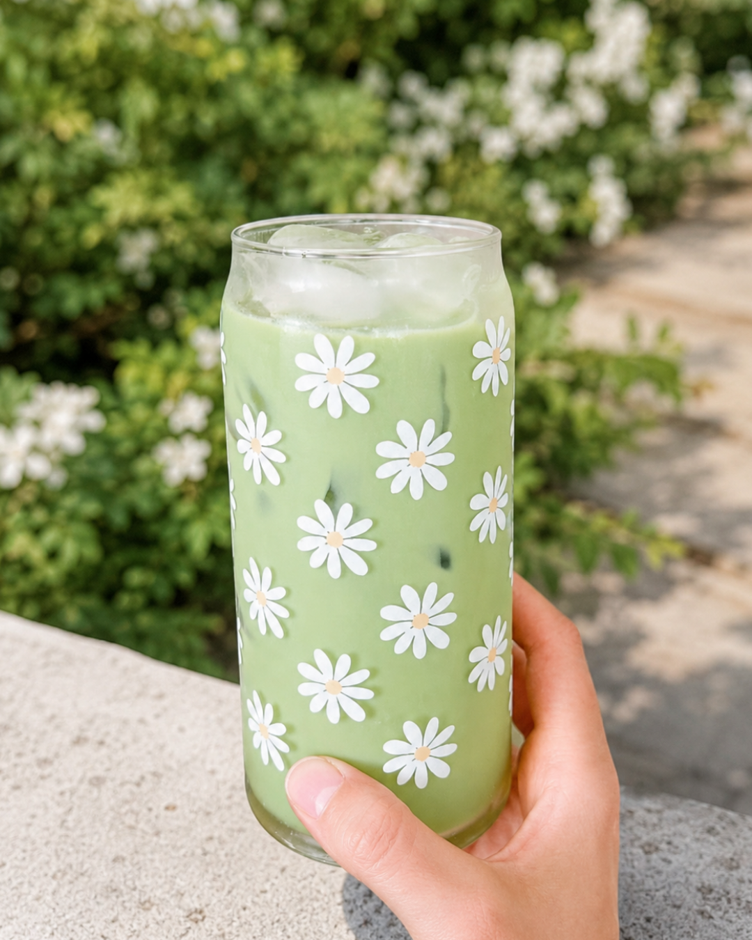 A hand holds a clear glass can-shaped tumbler decorated with white daisy flowers, filled with a light green iced drink, against a blurred outdoor garden background.