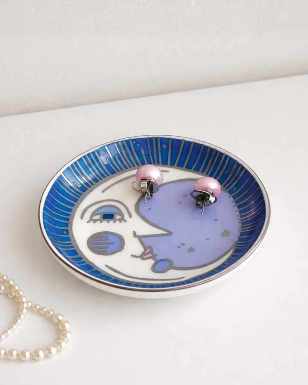 Moon face trinket dish with pearl necklace and pink earrings on white table, decorative celestial jewelry organizer tray