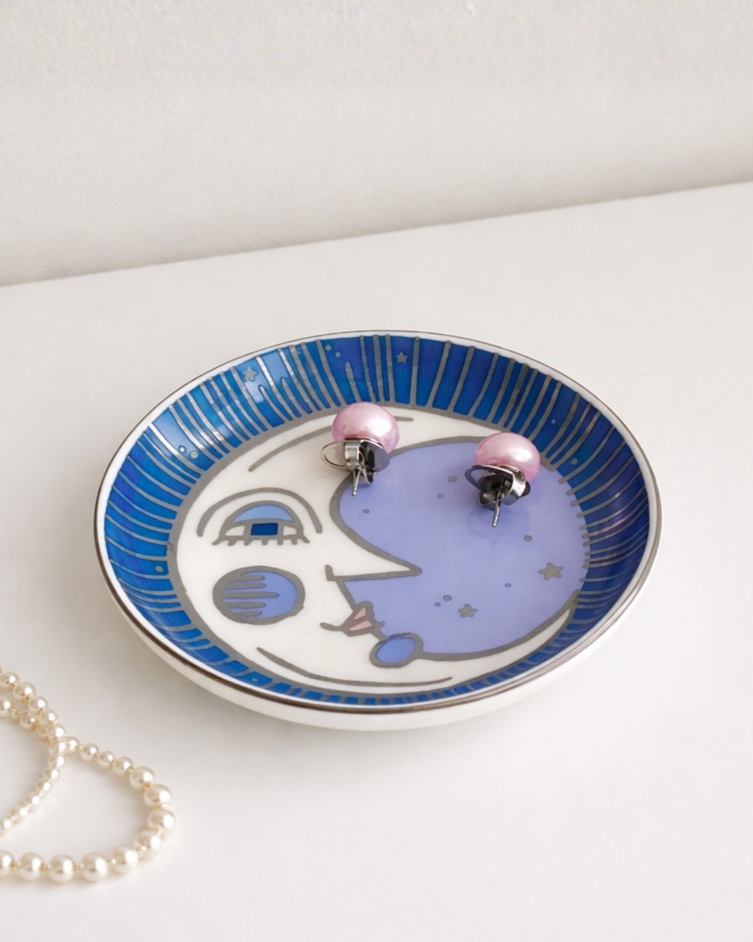 Moon face trinket dish with pearl necklace and pink earrings on white table, decorative celestial jewelry organizer tray