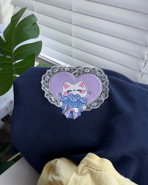 Close-up of navy blue sweatshirt showing heart-shaped lace cat patchwork embroidery design with purple bow, styled on a table.