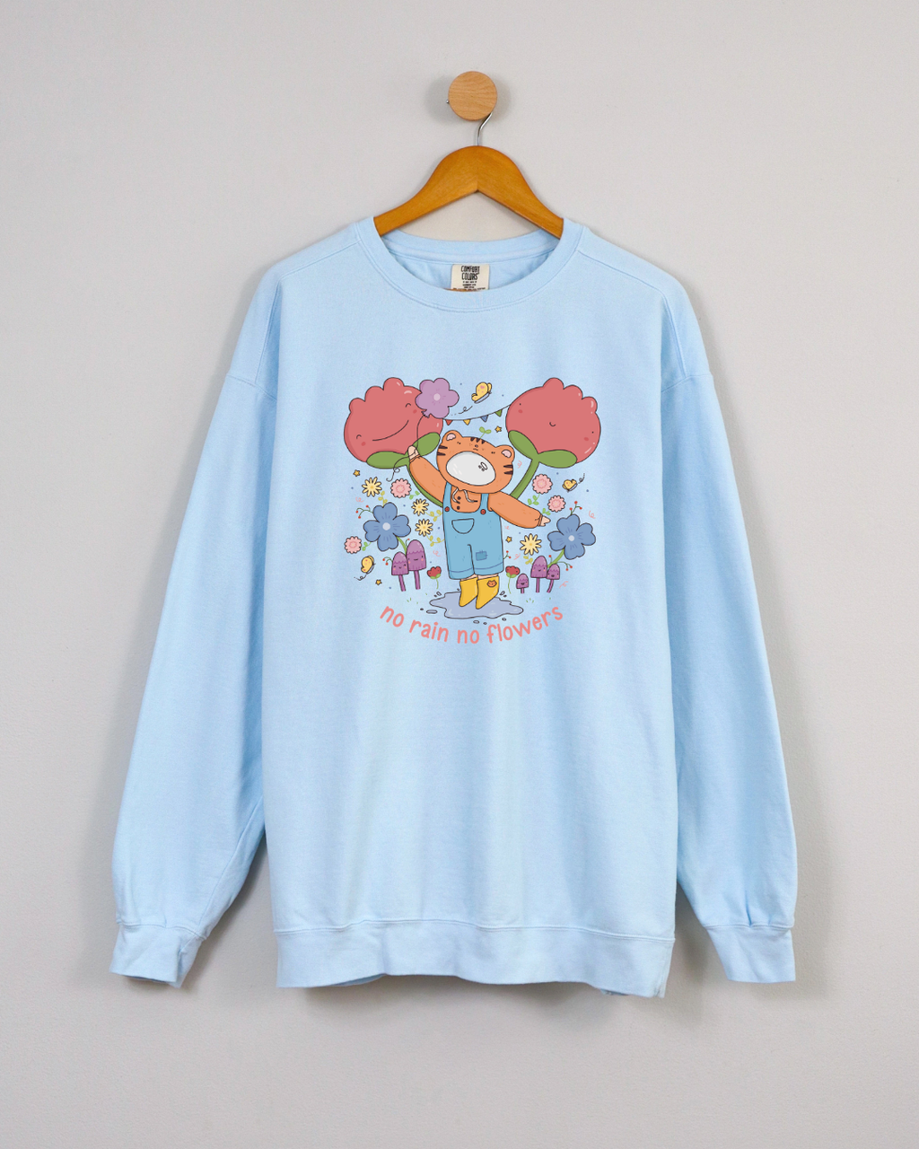 Sky blue crewneck sweatshirt with a playful illustration and the phrase “no rain no flowers,” featuring a storybook-style character surrounded by flowers