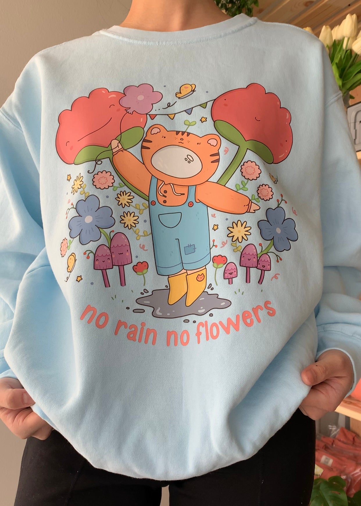 Model wearing a sky blue graphic crewneck sweatshirt featuring a playful illustrated character surrounded by flowers and the phrase “No rain, no flowers,” styled for a calm, comforting everyday look.