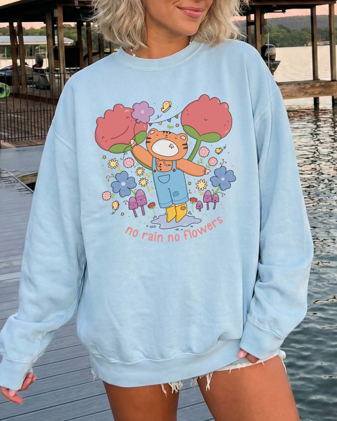 Woman wearing a sky blue “no rain no flowers” graphic sweatshirt outdoors by the water, styled as a relaxed, cozy everyday outfit