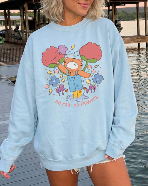 Woman wearing a sky blue “no rain no flowers” graphic sweatshirt outdoors by the water, styled as a relaxed, cozy everyday outfit