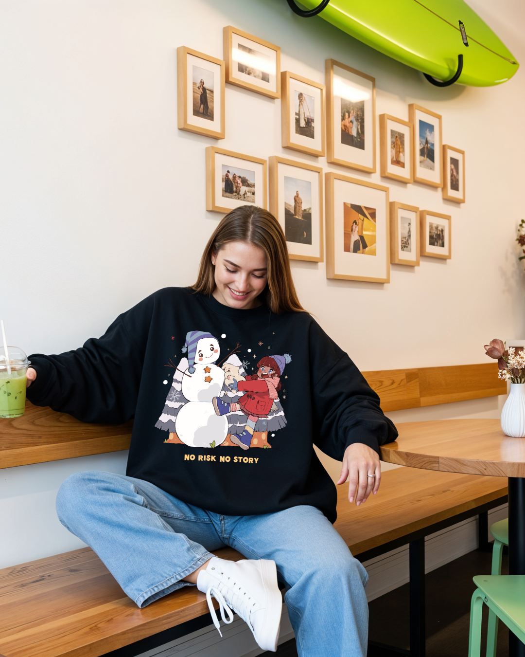Woman wearing a black “no risk no story” graphic crewneck sweatshirt with a nostalgic winter illustration, styled casually indoors