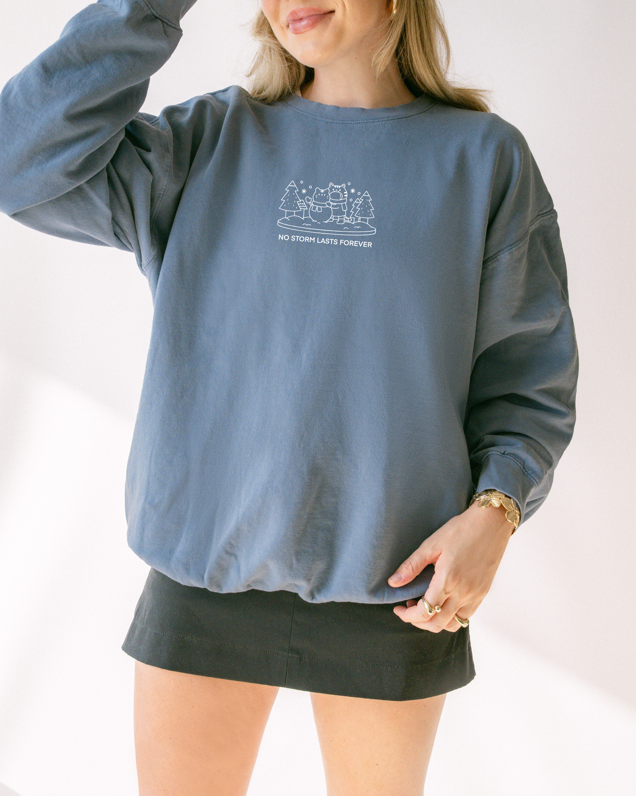 Model wearing a dusty blue embroidered sweatshirt with a winter scene and the text “No Storm Lasts Forever,” styled for calm everyday wear.
