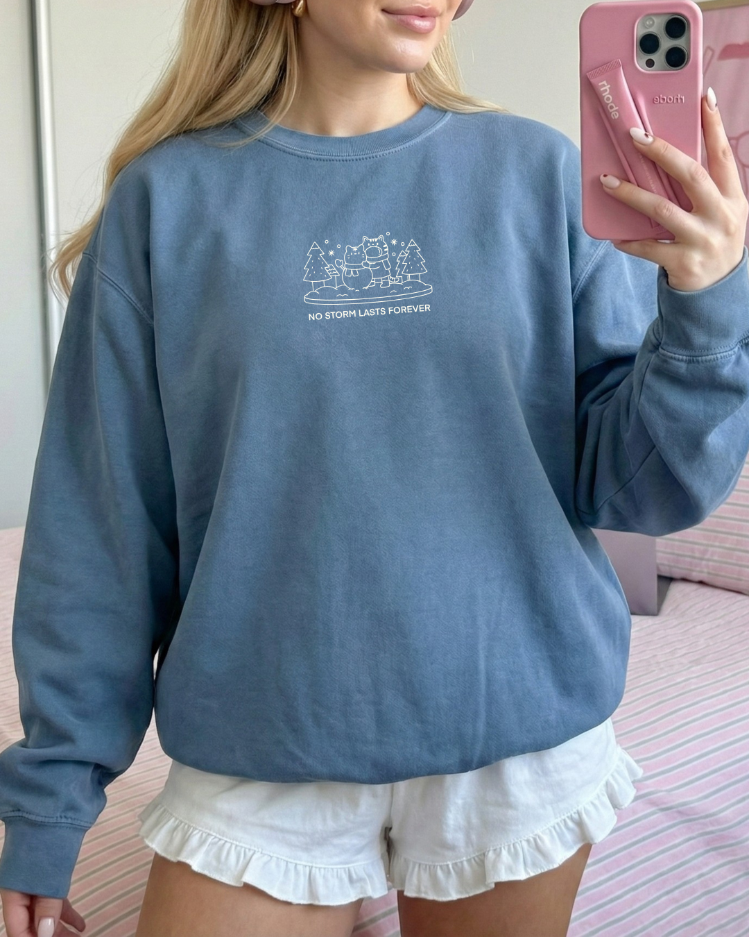 Dusty blue embroidered crewneck sweatshirt featuring a small winter scene and the phrase “No Storm Lasts Forever,” worn casually indoors