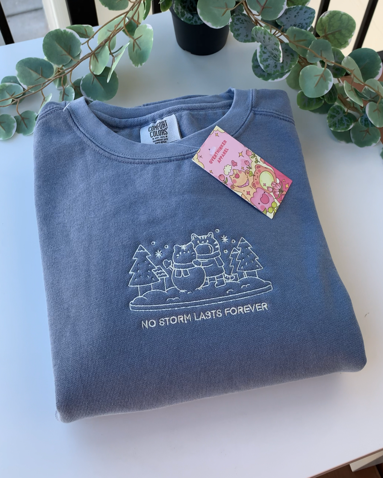 Folded dusty blue crewneck sweatshirt with subtle embroidery, styled with greenery for a calm, cozy aesthetic