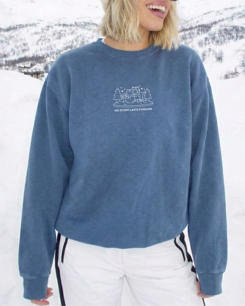 Person wearing a dusty blue “No Storm Lasts Forever” embroidered sweatshirt outdoors in a snowy setting, styled for comfort during winter weather.