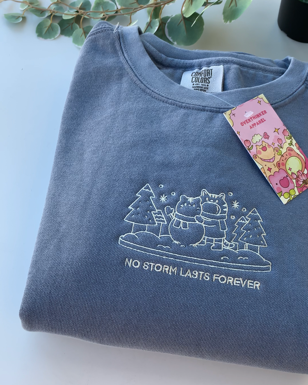 Close-up of white embroidered winter scene and text reading “No Storm Lasts Forever” on a dusty blue sweatshirt