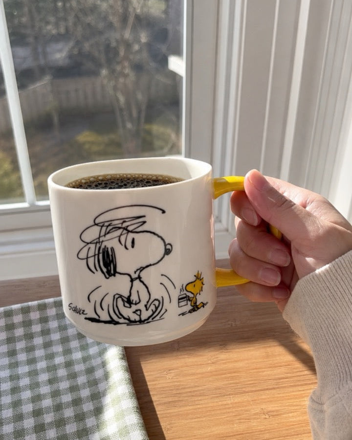 A hand holds a yellow-handled Snoopy mug featuring a disheveled Snoopy and Woodstock with a tiny coffee cup, near a sunlit window with a checkered cloth on a wooden surface.