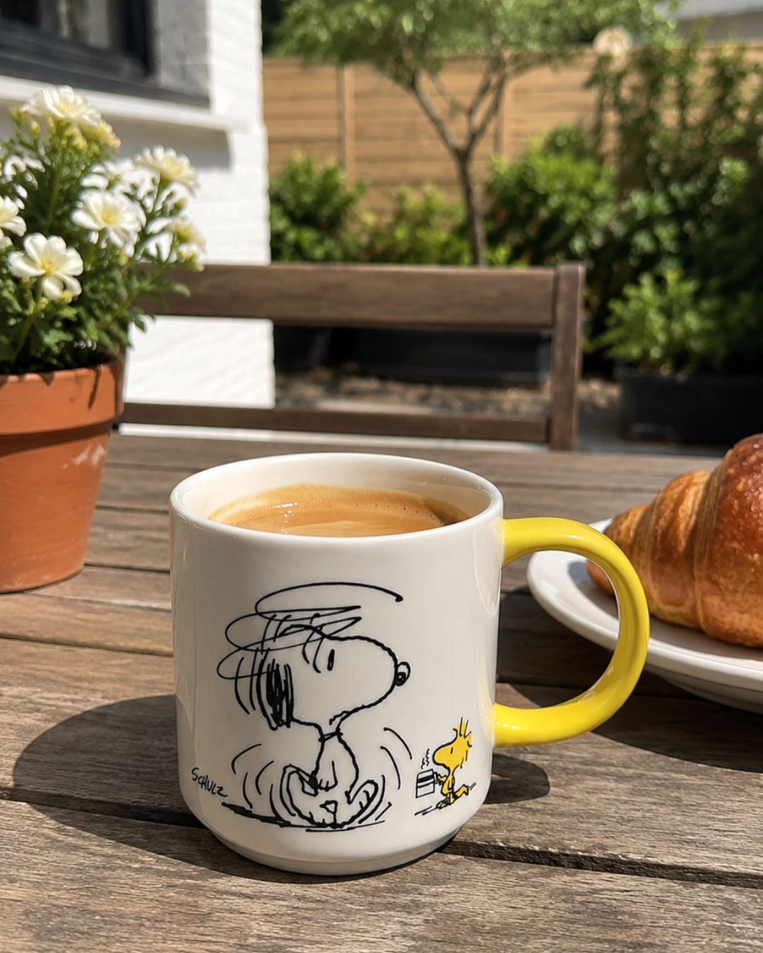 A yellow-handled Snoopy mug featuring a disheveled Snoopy shaking off sleep with Woodstock holding a tiny coffee cup, styled on an outdoor patio table with a croissant and flowers.