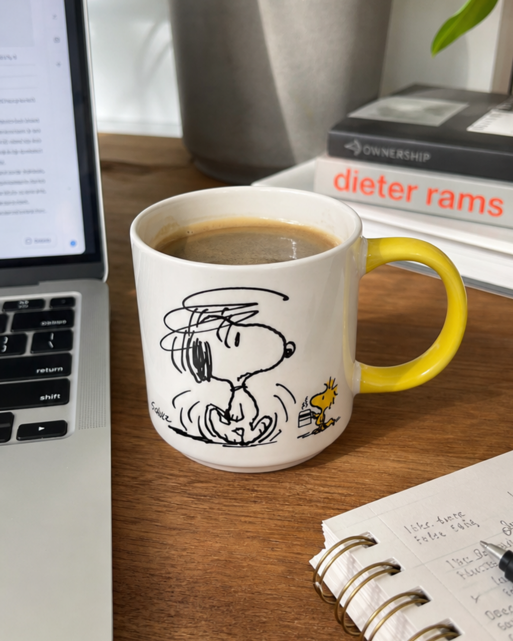A yellow-handled Snoopy mug featuring a disheveled, grumpy Snoopy shaking off sleep with Woodstock walking by holding a tiny cup of coffee, styled on a desk with a laptop, books, and a notebook.