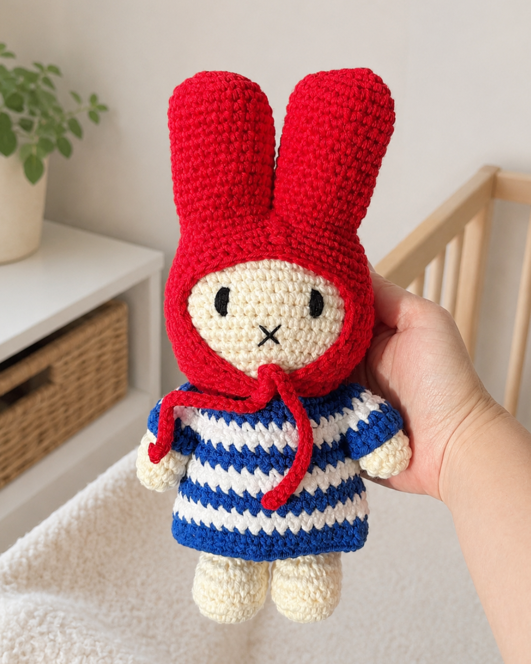 A handmade crocheted doll with long red bunny ears, a cream face, and a blue and white striped outfit, held in a person's hand against a soft indoor background.