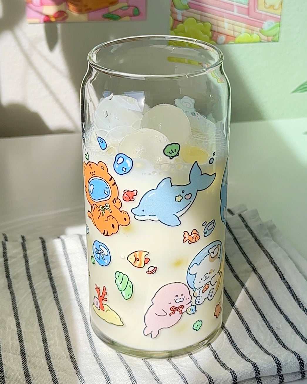 Clear 20 oz glass can cup with cute ocean-themed illustrations, shown filled with ice and a light drink on a cozy kitchen surface.
