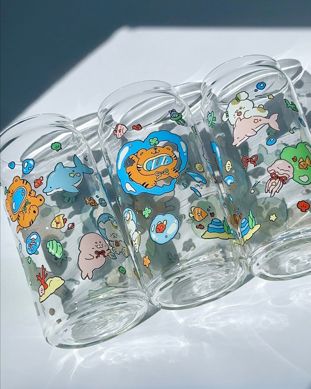 Three clear 20 oz glass can cups featuring a cute ocean playground illustration with playful underwater characters, styled together in natural light.