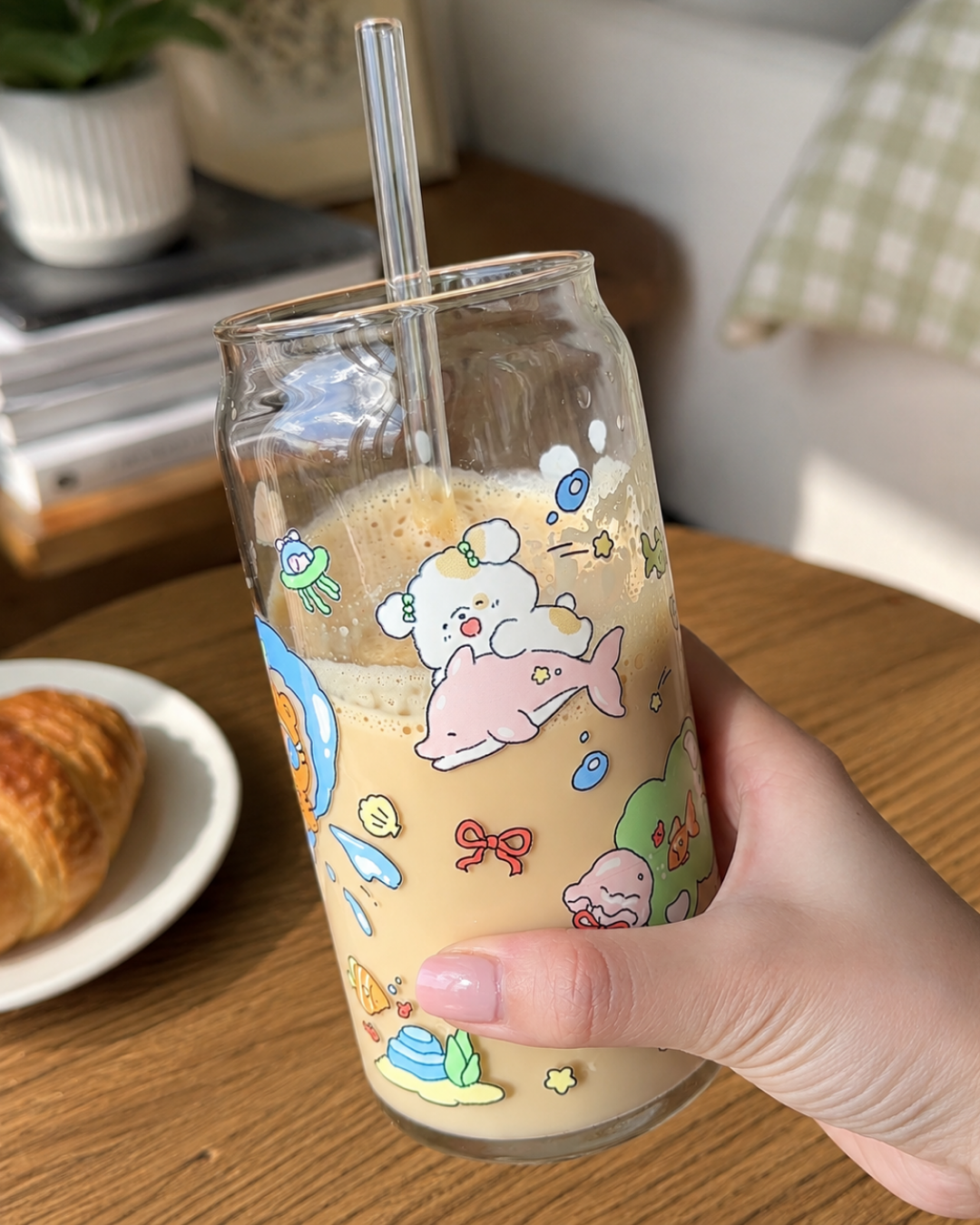 A hand holding a clear glass can-style tumbler with a straw, decorated with colorful cartoon sea creatures and a white bear riding a pink dolphin. The glass is filled with a light tan iced drink and sits on a wooden table with a croissant in the background.