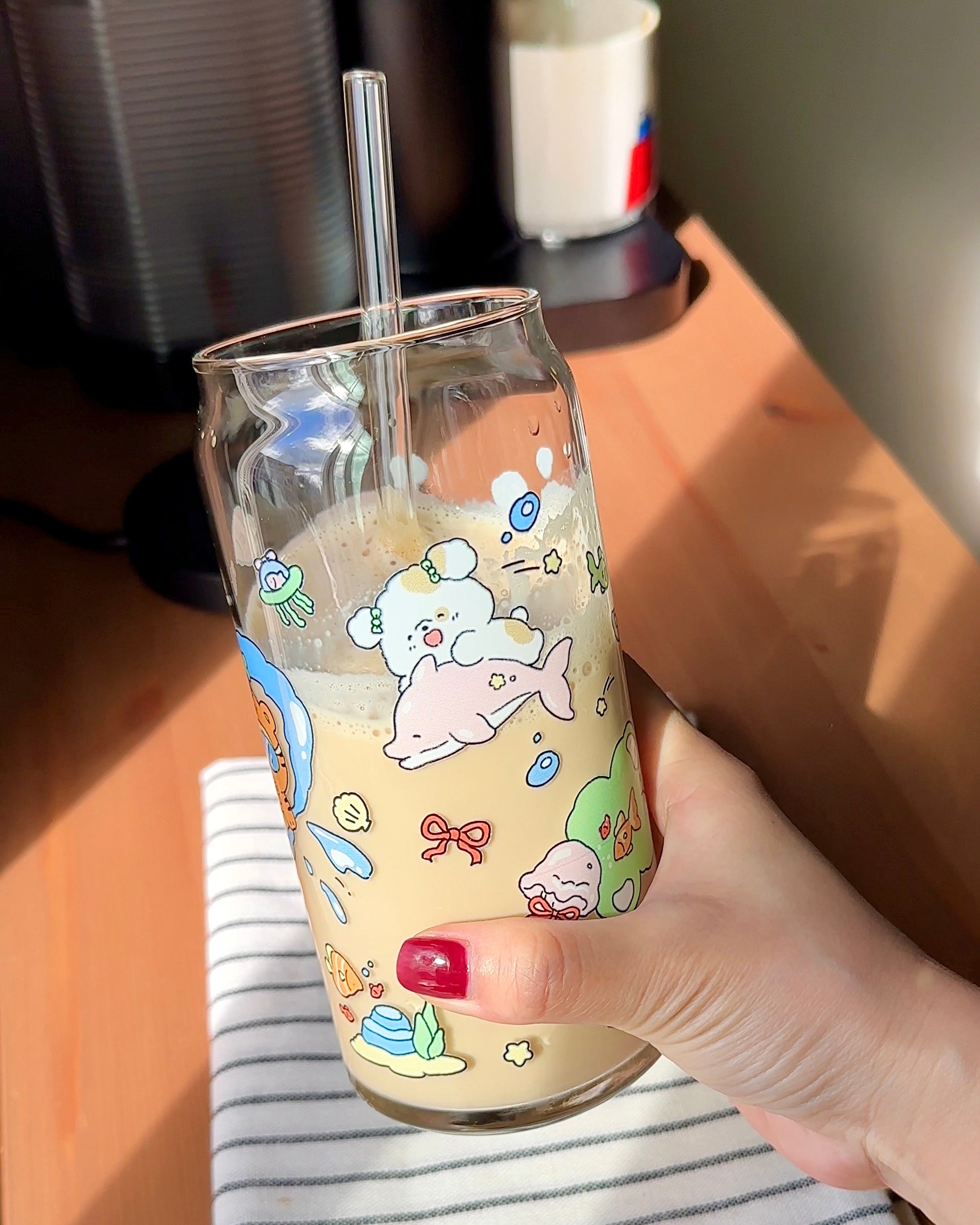 Hand holding a 20 oz ocean playground glass can filled with iced coffee, featuring soft pastel underwater characters and a reusable glass straw.