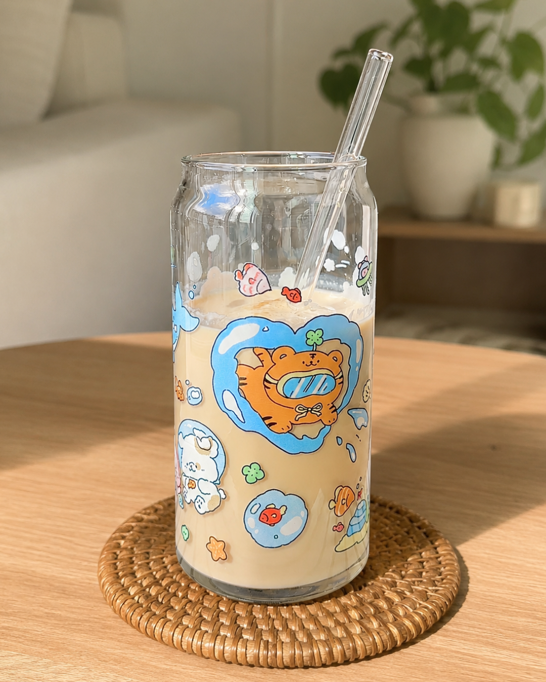 A clear glass can-shaped tumbler with a cartoon tiger illustration and small colorful doodles, filled with a light beige drink and a clear straw, sitting on a woven round coaster on a wooden table.