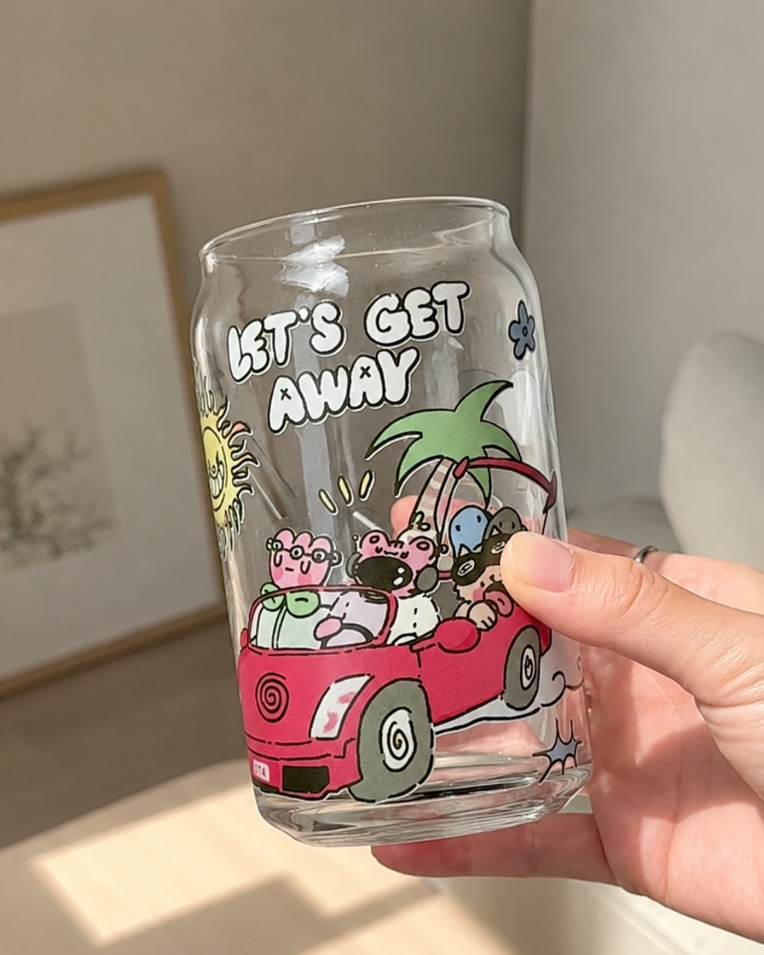 A hand holding a clear glass can-shaped tumbler with a colorful cartoon illustration of characters in a red car and the text 'LET'S GET AWAY'.