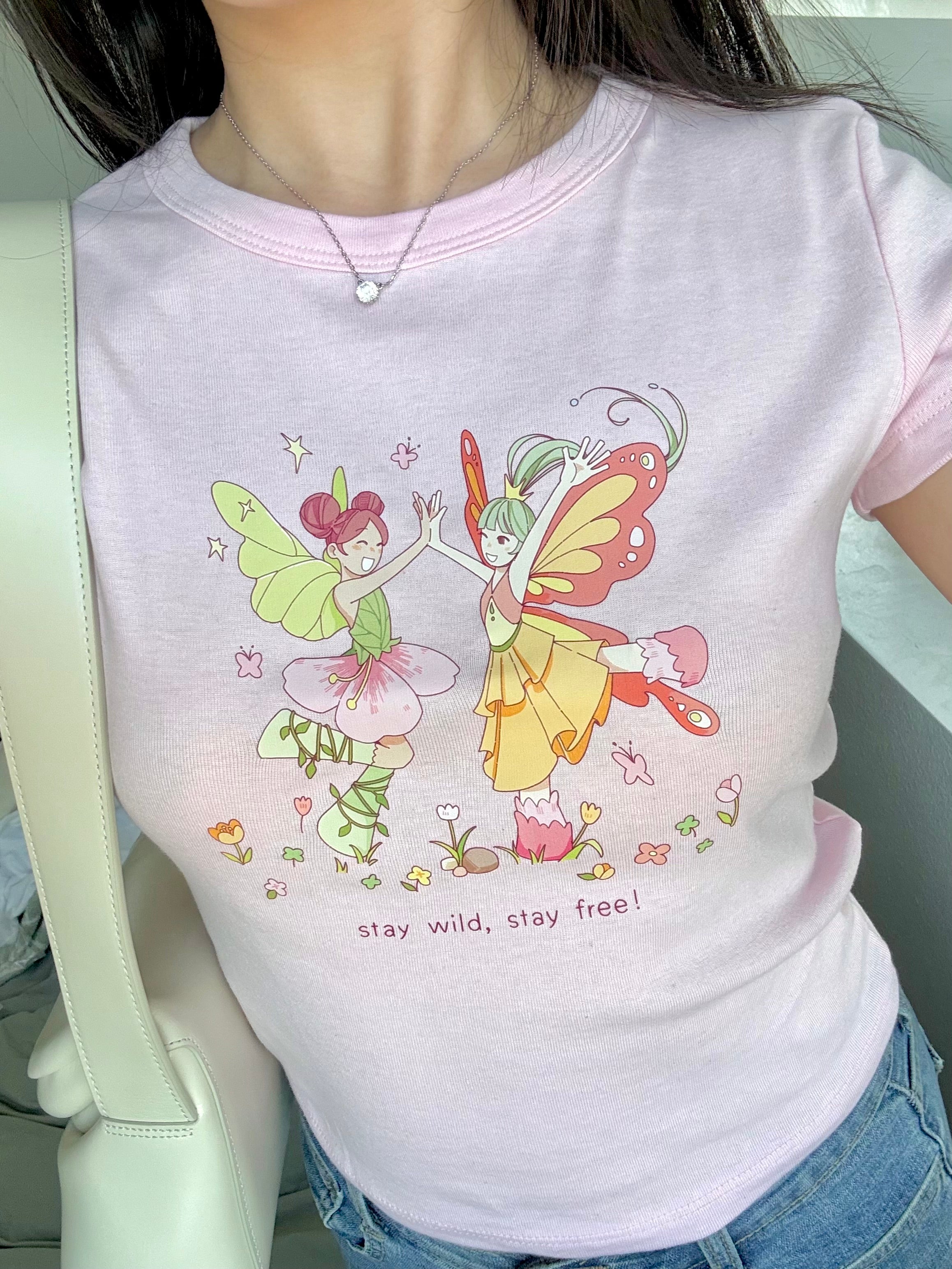 Pastel pink fairycore baby tee featuring two dancing fairies and the phrase “stay wild, stay free”