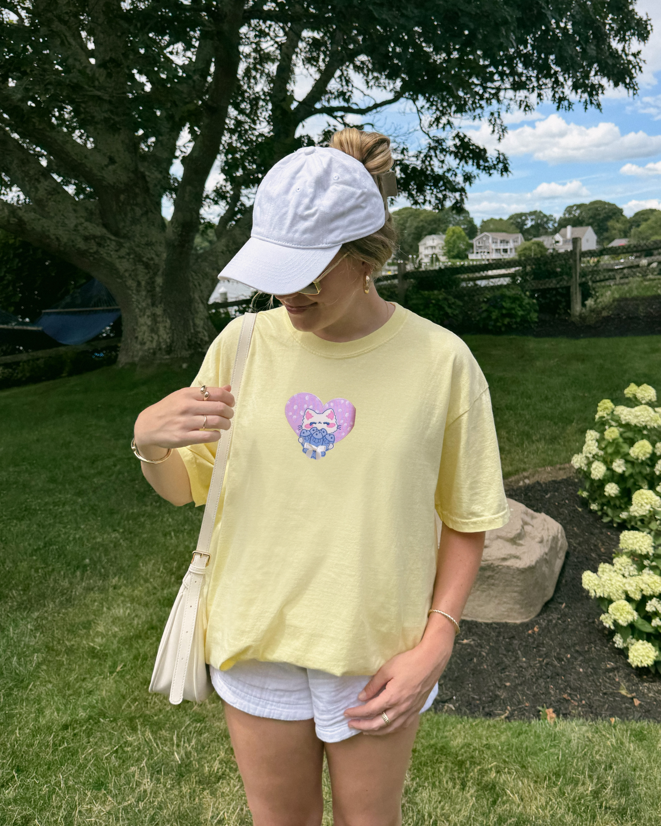 Woman wearing a pastel yellow Comfort Colors t-shirt with an embroidered white cat patch inside a lilac heart, styled casually outdoors in a garden.