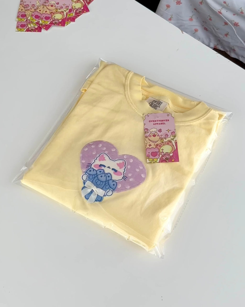 Folded pastel yellow Comfort Colors t-shirt with embroidered cat patch, packaged with Overthinker Apparel tag on a white surface.