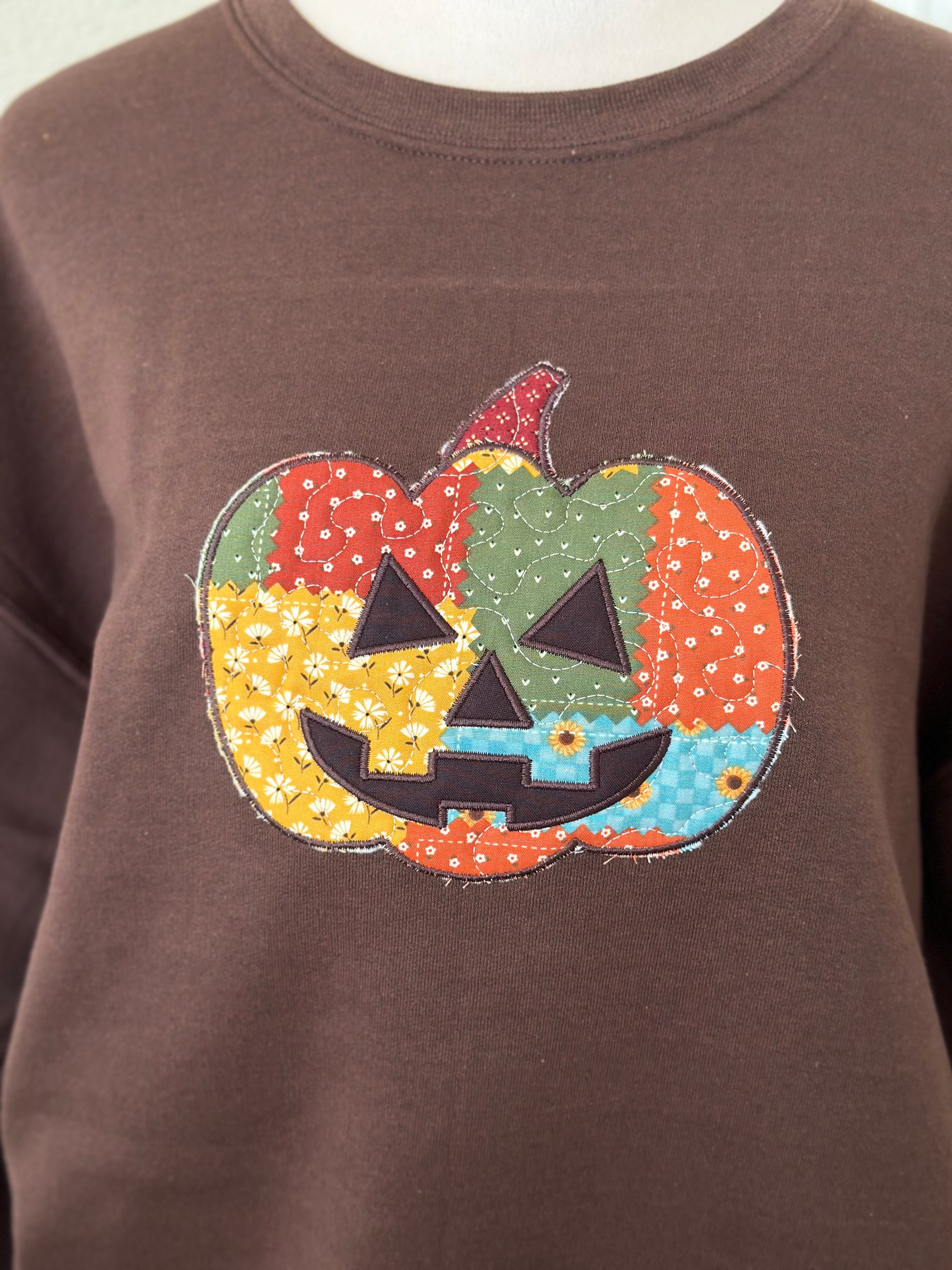 Close-up of a brown sweatshirt featuring a colorful patchwork jack-o’-lantern applique with stitched details.
