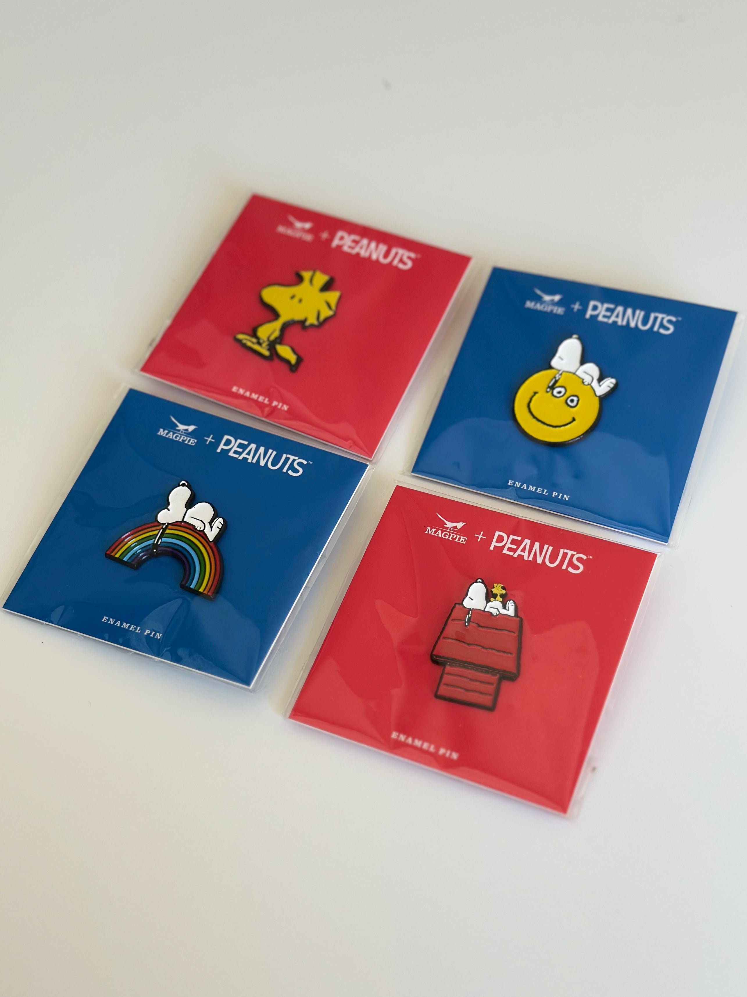 Set of four Peanuts enamel pins featuring Woodstock, Snoopy on rainbow, Snoopy smiley face, and Snoopy with Woodstock on doghouse.