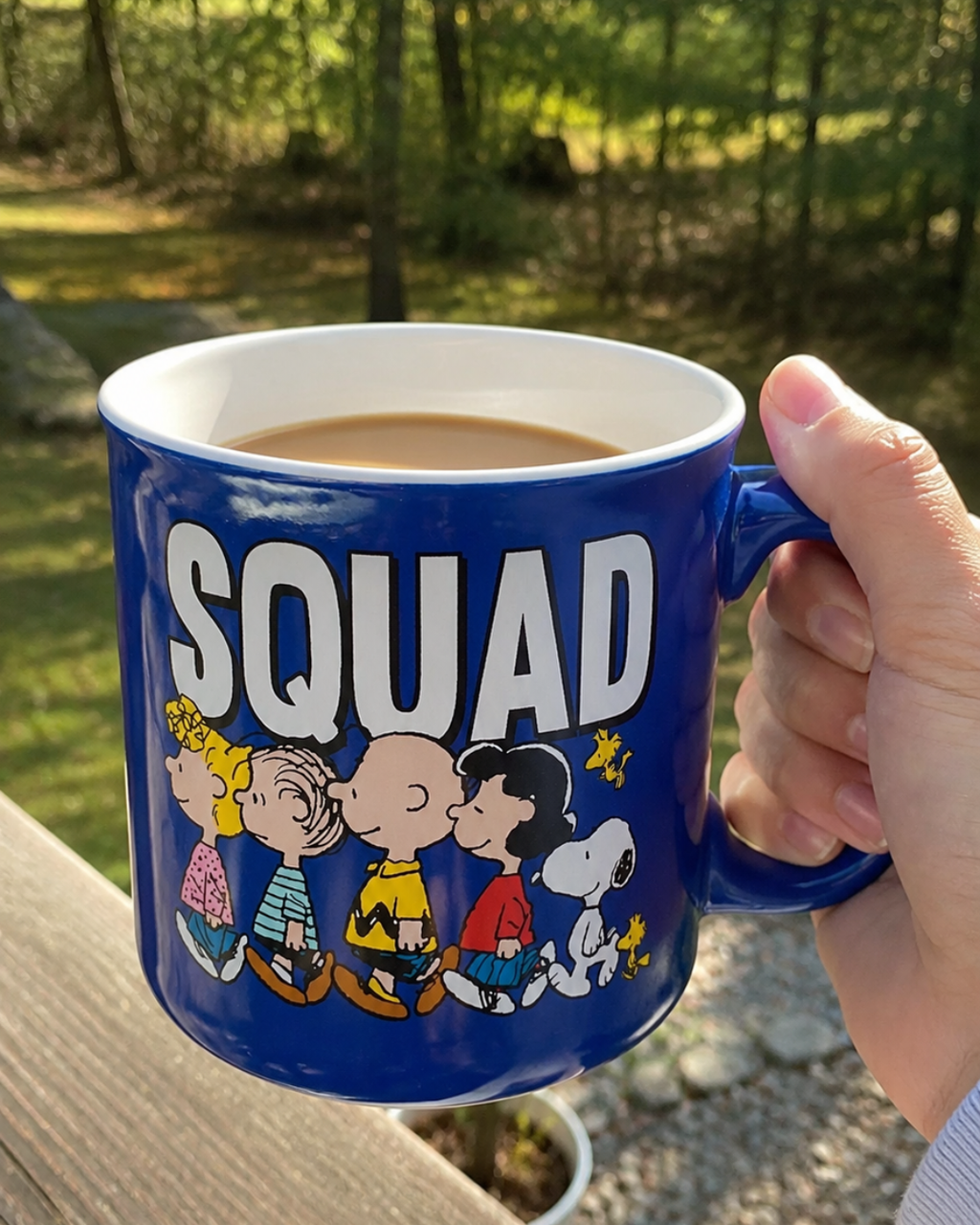 A hand holds a 20 oz cobalt blue Peanuts Squad mug outdoors on a wooden deck railing, with the Snoopy and Peanuts gang design visible and a forest backdrop.