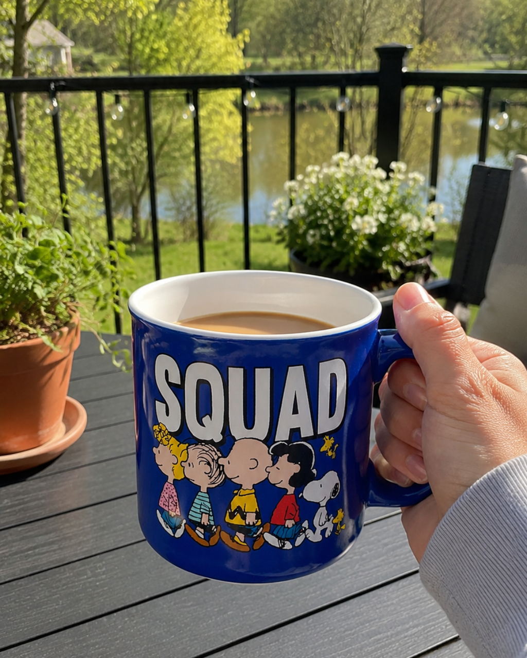 A hand holds an oversized Snoopy Peanuts Squad coffee mug outdoors on a patio table, with string lights, potted flowers, and a pond visible in the background.
