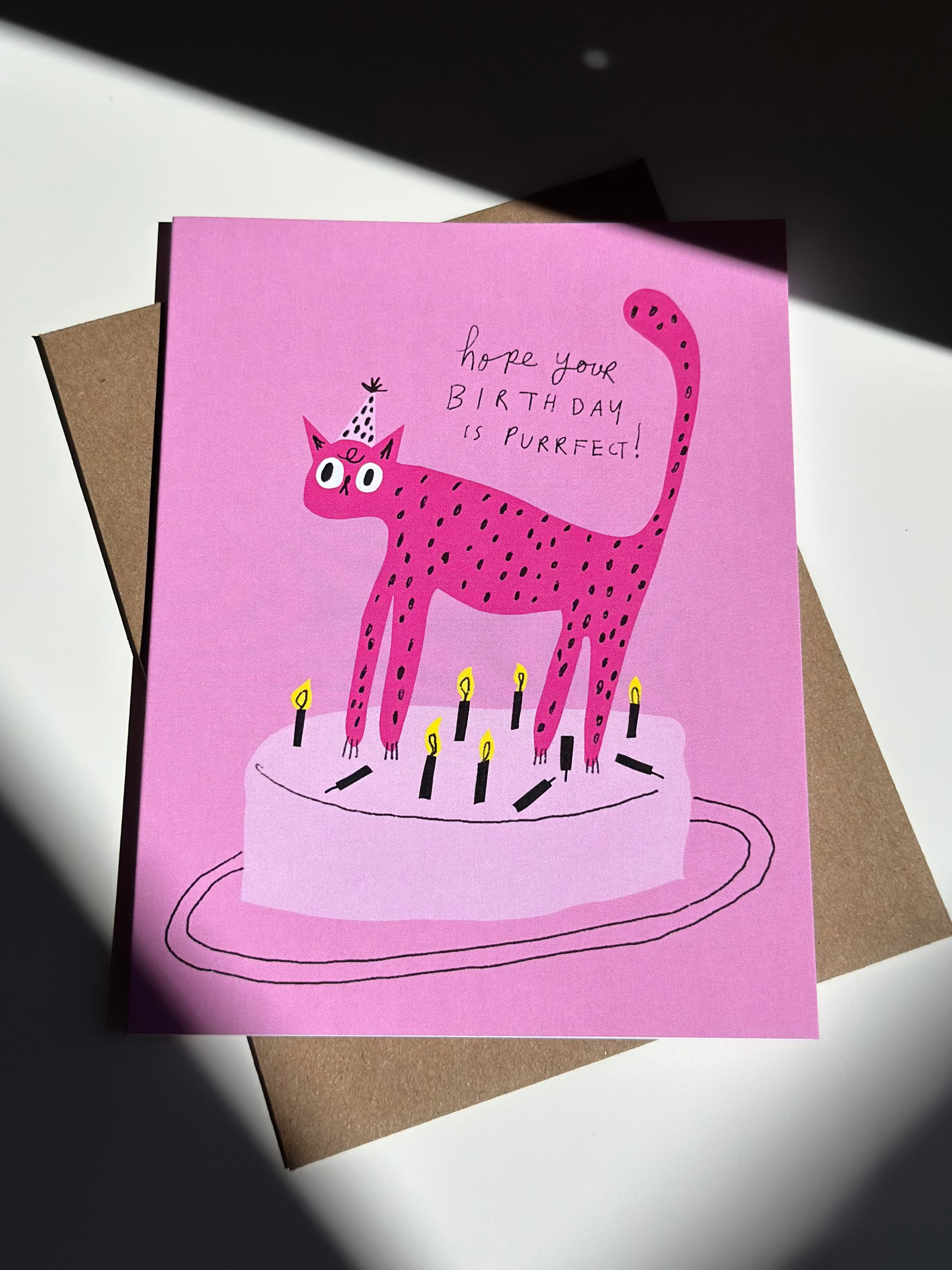 Pink cat birthday card with cartoon spotted cat wearing a party hat standing on a birthday cake, surrounded by lit candles, and text that reads ‘Hope your birthday is purrfect!’ on pink background with brown kraft envelope.