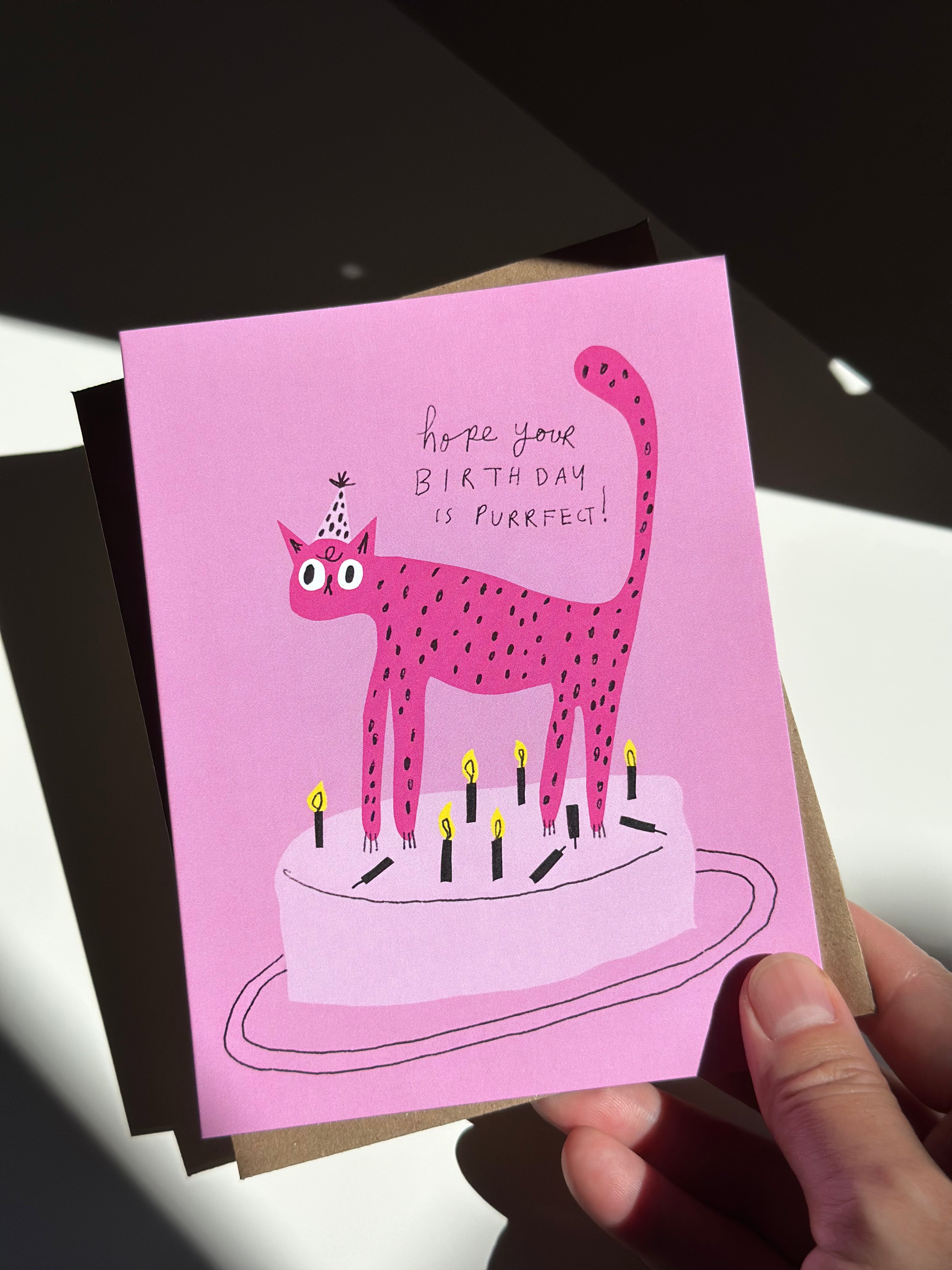 Pink cat birthday card with cartoon spotted cat wearing a party hat standing on a birthday cake, surrounded by lit candles, and text that reads ‘Hope your birthday is purrfect!’ on pink background with brown kraft envelope.