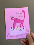 Pink cat birthday card with cartoon spotted cat wearing a party hat standing on a birthday cake, surrounded by lit candles, and text that reads ‘Hope your birthday is purrfect!’ on pink background with brown kraft envelope.
