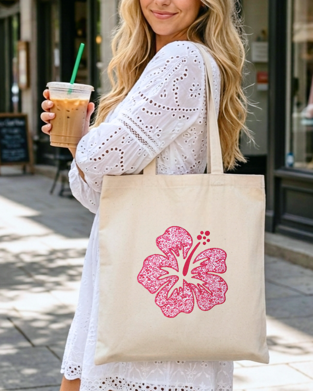 Woman carrying natural canvas tote with pink hibiscus embroidery while holding iced coffee, casual everyday outfit floral tote bag