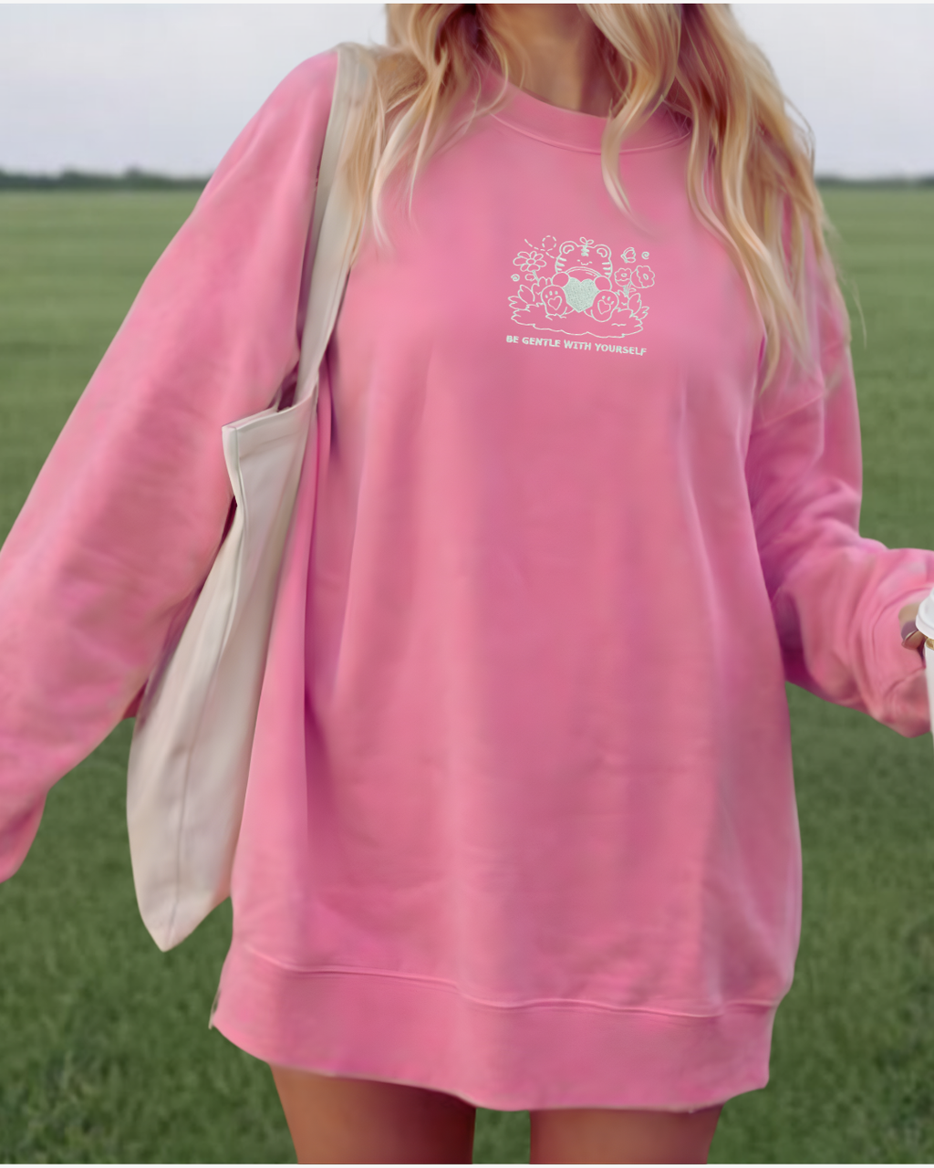 Woman wearing oversized pink crewneck sweatshirt with small white “Be Gentle With Yourself” bear graphic, cozy affirmation sweatshirt styled as casual everyday outfit.