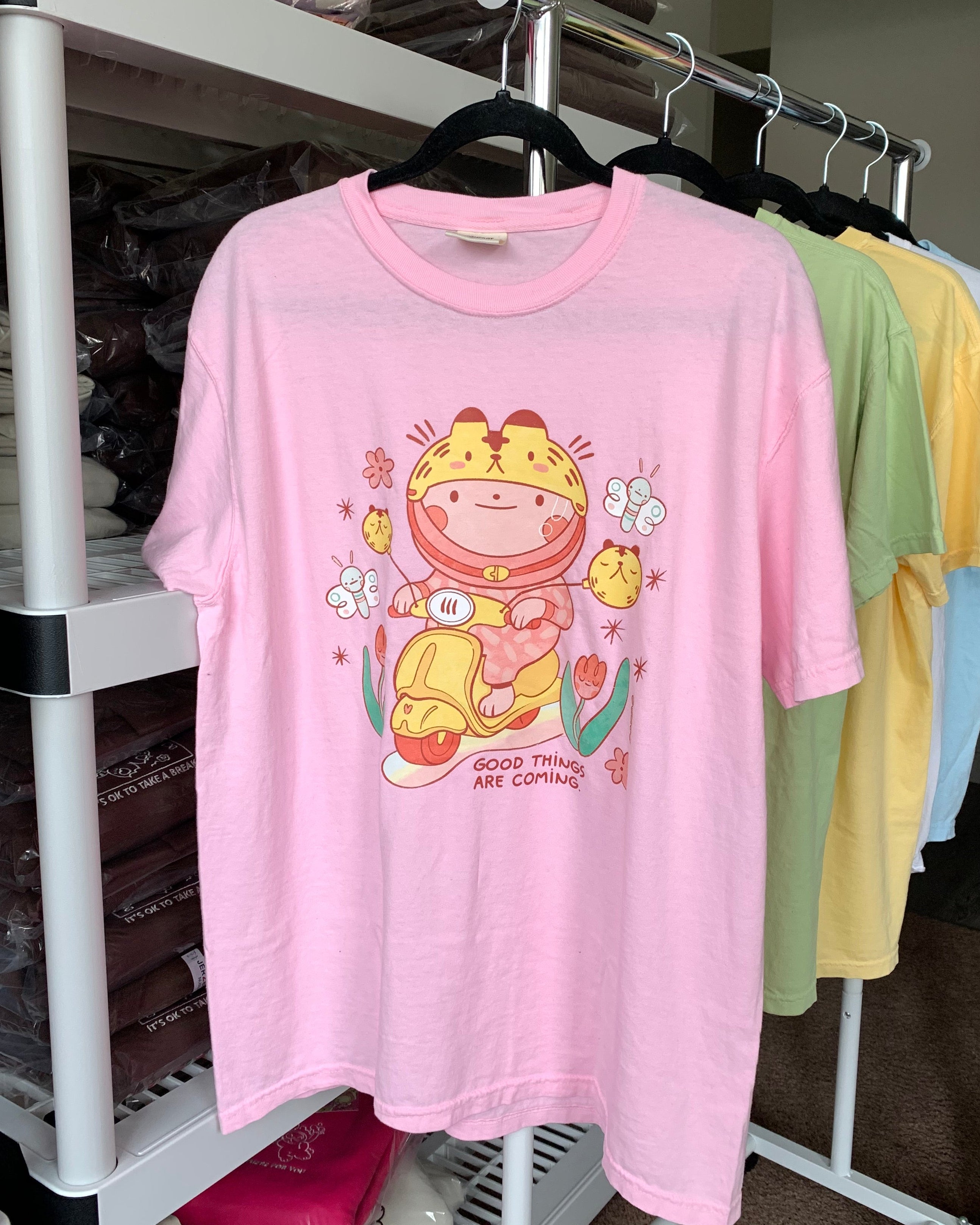 Pink graphic tee with cheerful illustration and hopeful good things are coming message hanging on clothing rack