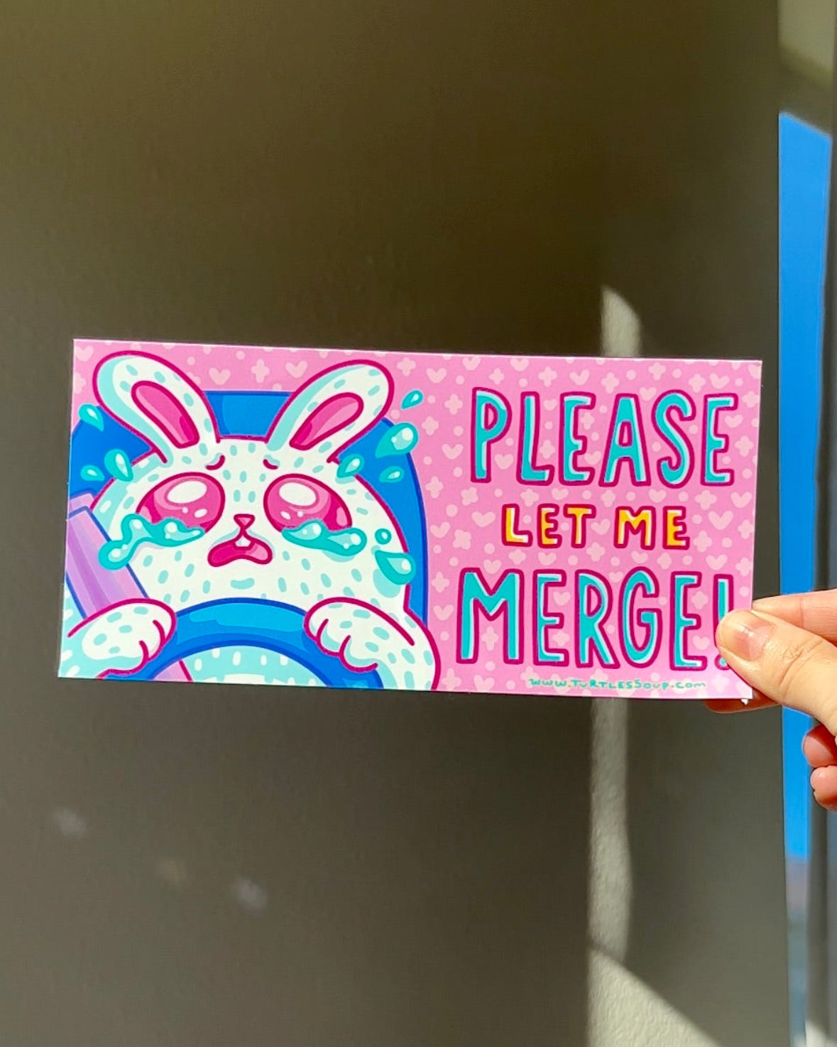 Hand holding a cute vinyl car sticker with a crying bunny and the words “Please Let Me Merge,” funny anxious driver decal for car windows.