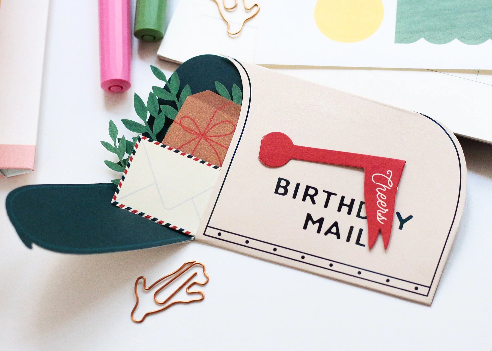 Whimsical pop-up birthday card shaped like a mailbox with “Cheers” flag, filled with 3D paper gifts, greenery, and a tiny envelope. A sweet and thoughtful birthday greeting card for friends or loved ones.

