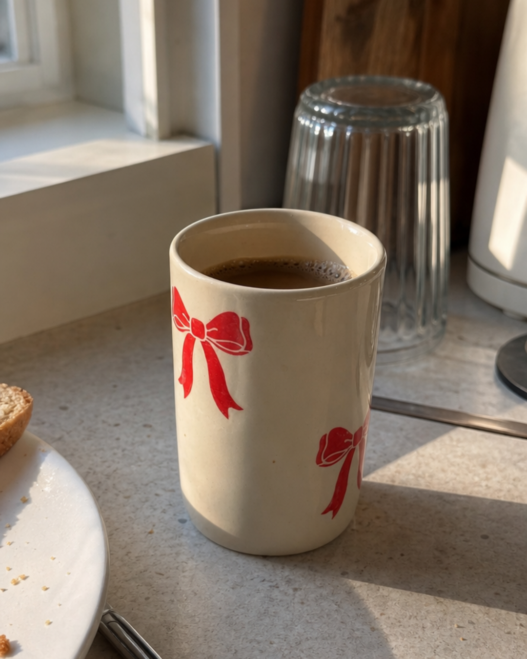 A red bow hand-painted ceramic mug filled with coffee sitting on a sunlit kitchen counter next to a pastry plate.