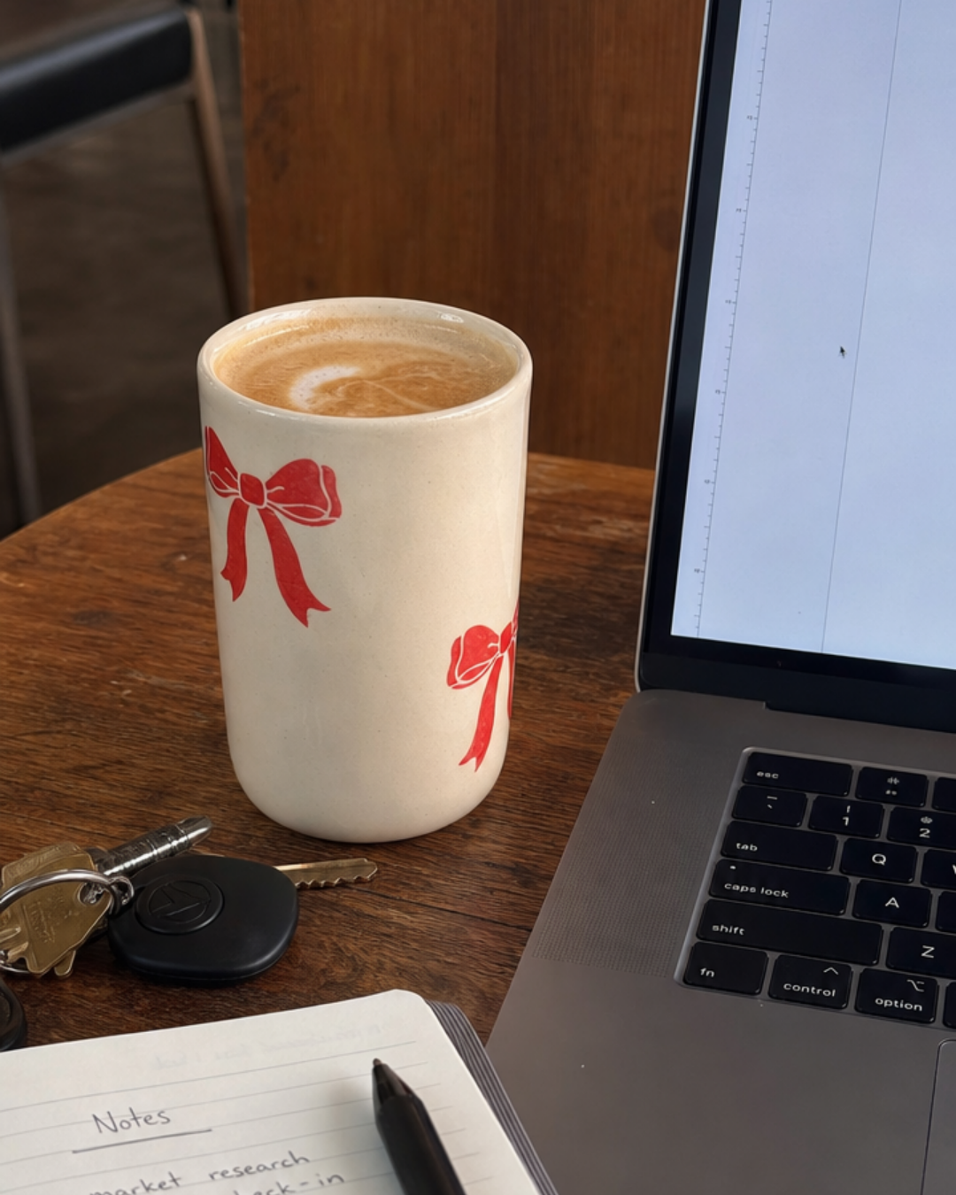 A red bow ceramic mug on a wood desk next to an open laptop, notebook, and keys, styled as a work-from-home coffee setup.