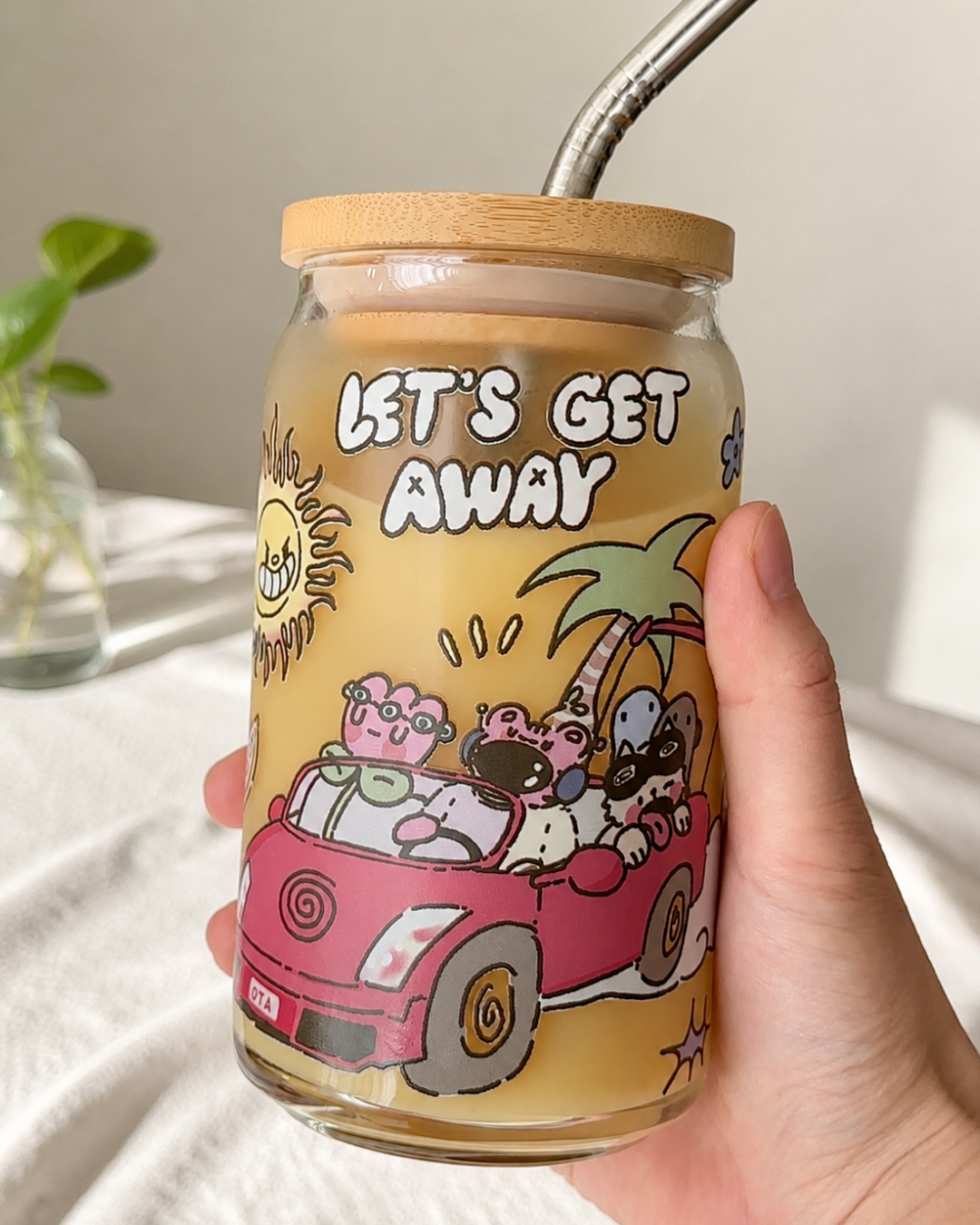 A hand holding a clear glass jar tumbler with a bamboo lid and metal straw, filled with a yellow drink. The tumbler features a cartoon illustration of characters in a pink car with the text 'LET'S GET AWAY'.