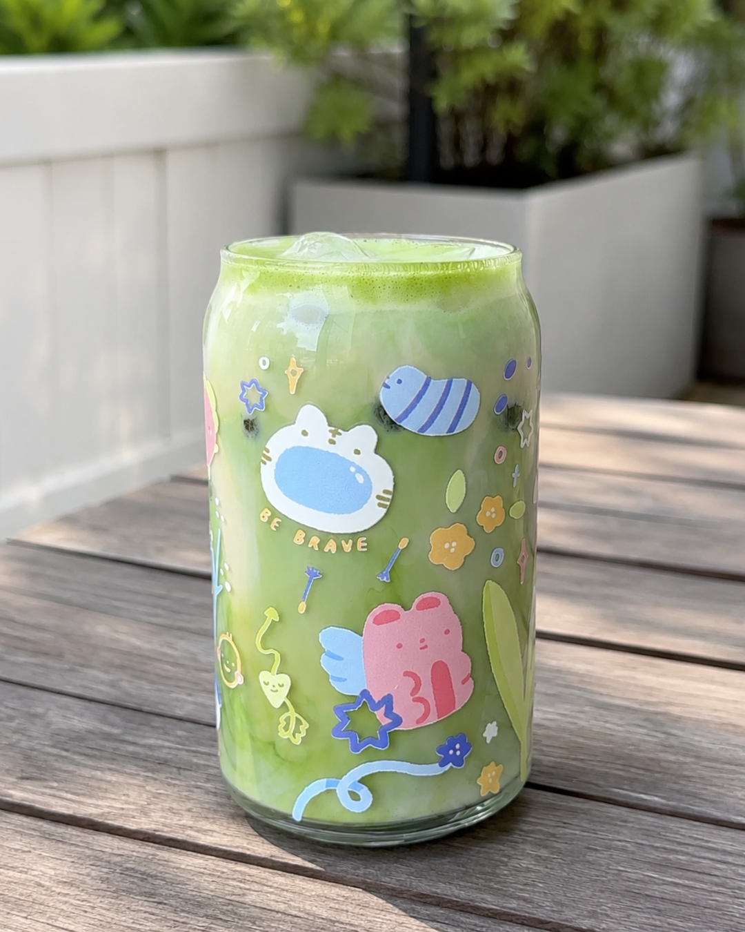 A clear glass can-shaped tumbler filled with a matcha drink, decorated with cute cartoon characters including a blue bee, a pink winged bear, and a white cat face, sitting on a wooden table outdoors.