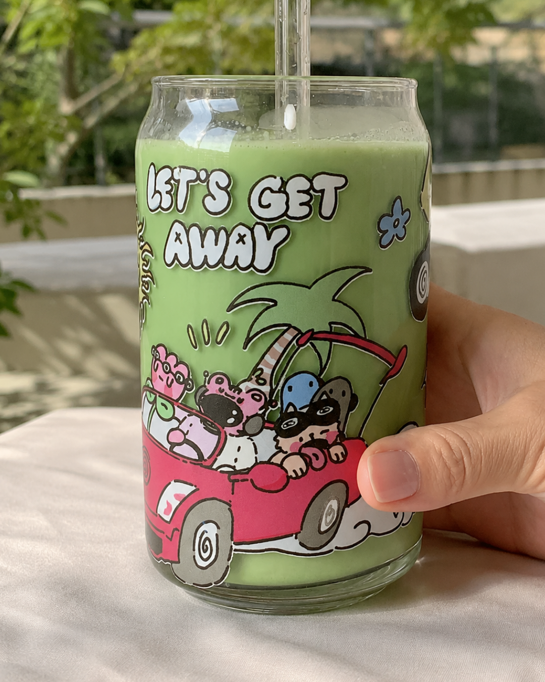 A hand holding a clear glass can-shaped tumbler with a straw, filled with a light green drink. The can features a cartoon illustration of characters in a red car with the text 'LET'S GET AWAY' on a light green background.