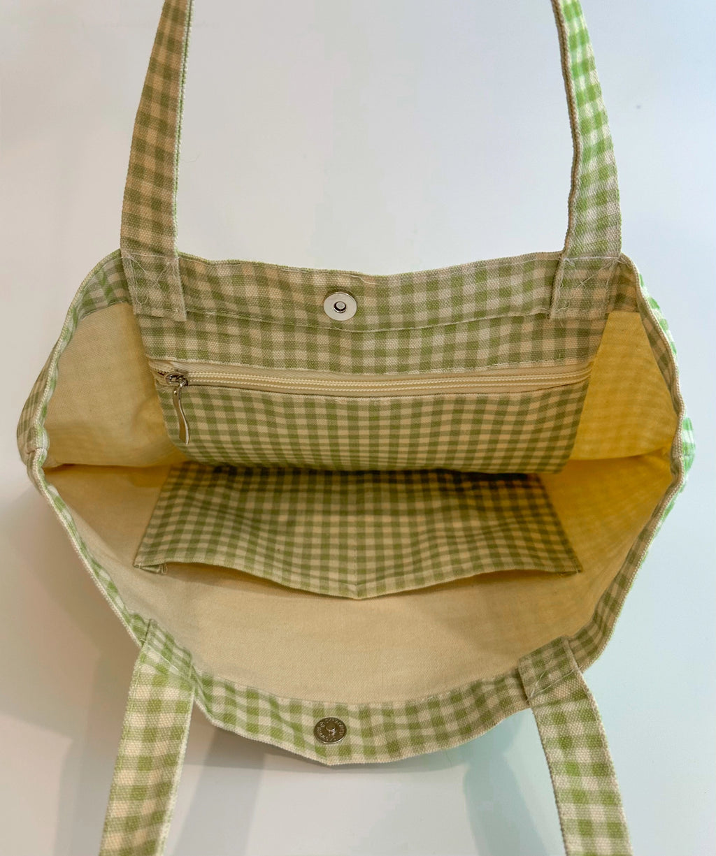 Inside view of a sage gingham canvas tote bag showing zip pocket, slip pocket, and magnetic snap closure for everyday organization.