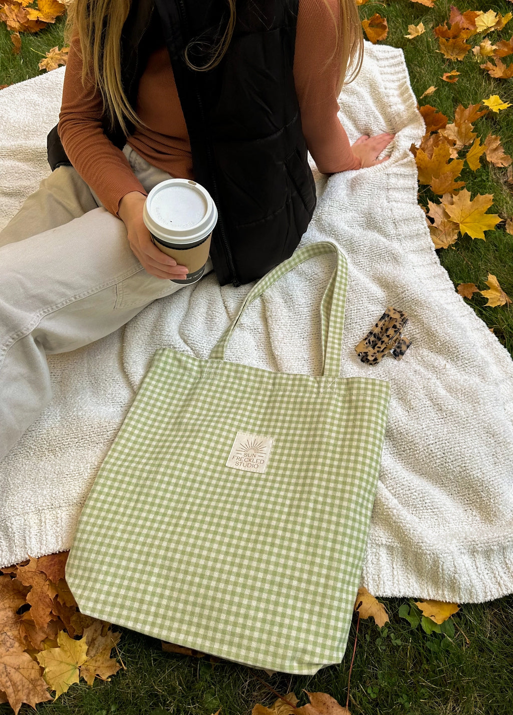 Sage green gingham canvas tote bag resting on a blanket outdoors, lightweight everyday tote styled for coffee runs and farmers markets.