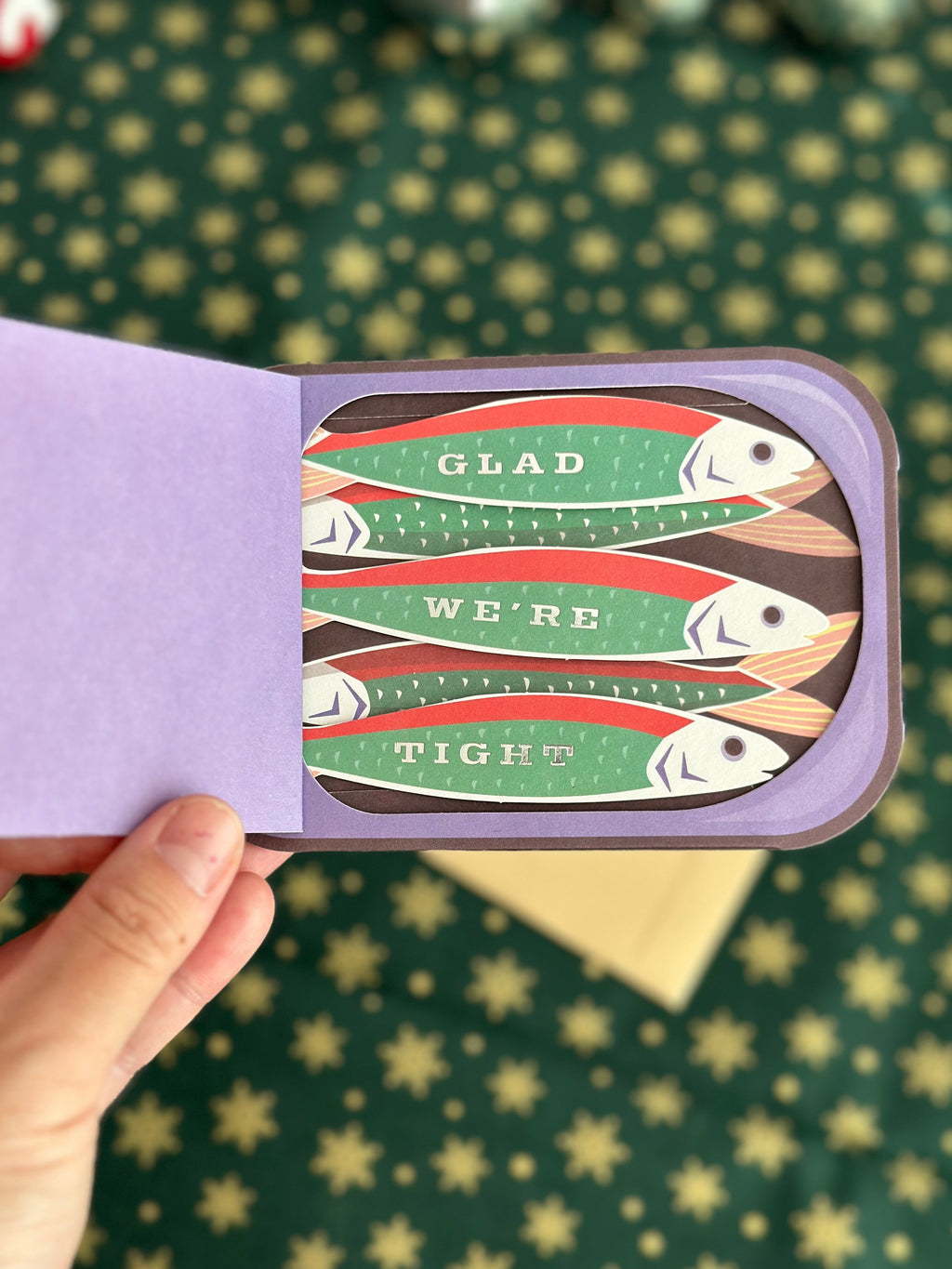 Hand holding the open sardine-can card to show the message “GLAD WE’RE TIGHT” on the fish design.