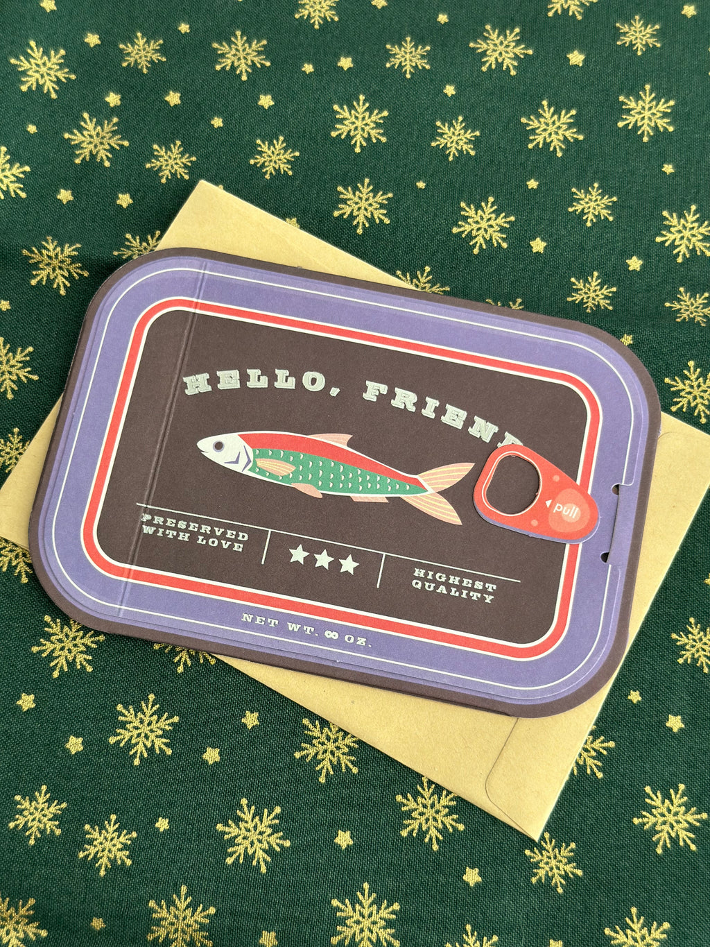 Sardine-can style holiday greeting card on green fabric with gold snowflakes, cover reads “HELLO, FRIEND” with pull tab and fish illustration.