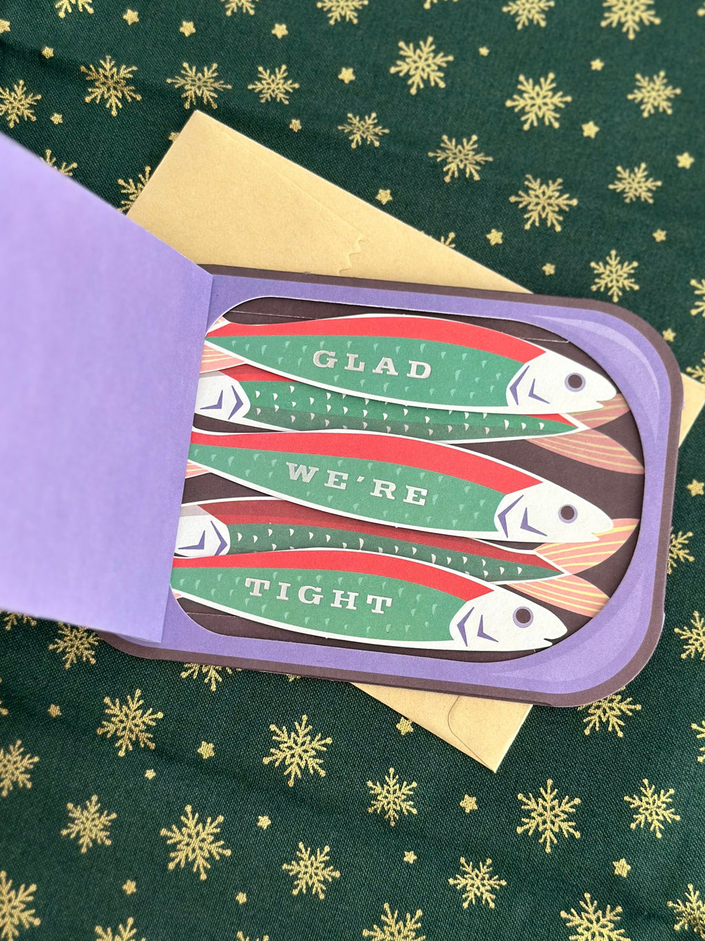 Inside of sardine-can card showing three fish with the message “GLAD WE’RE TIGHT” against a purple flap and gold snowflake background.